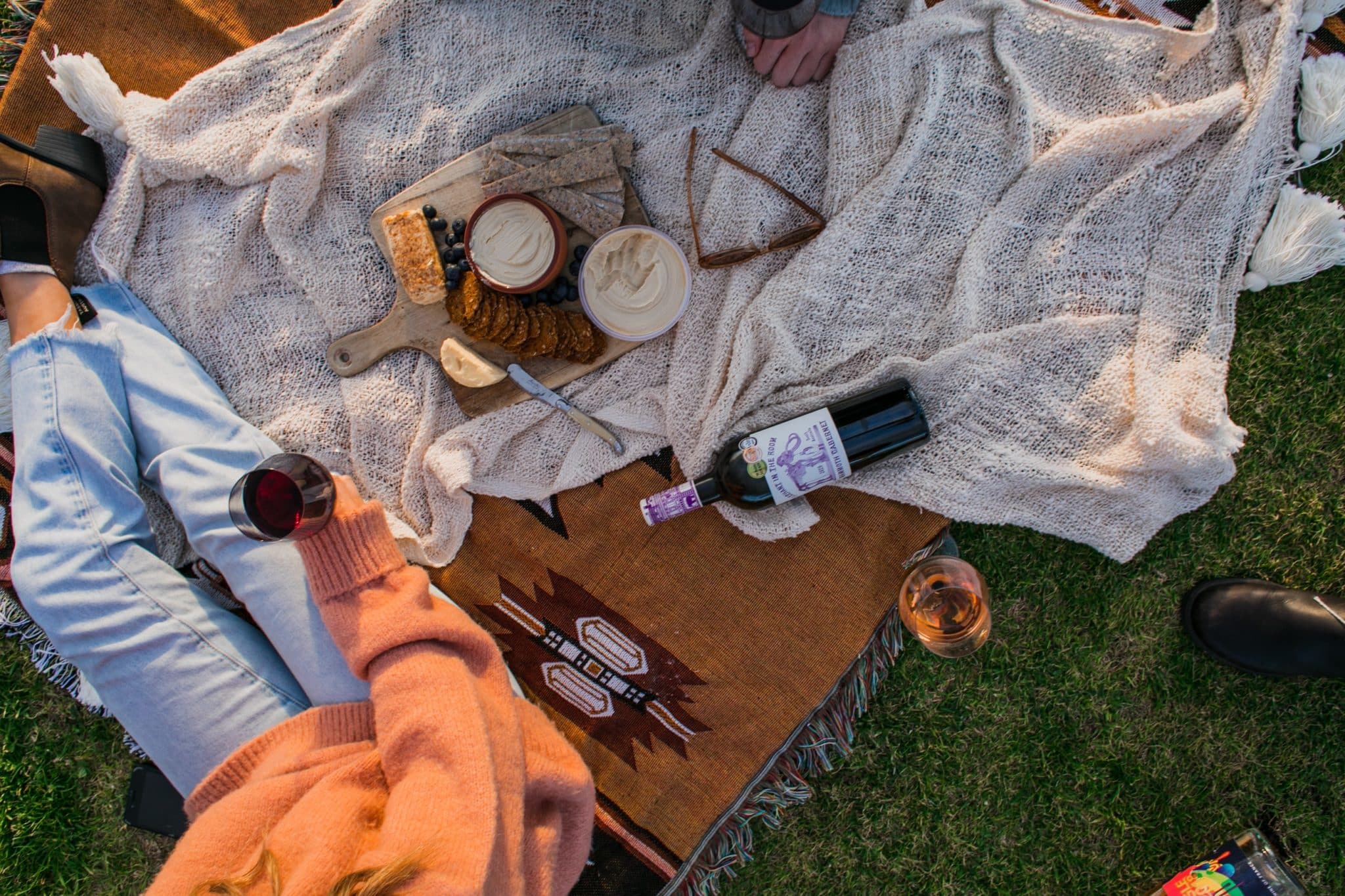 picnic con vino, humus, tostadas y fruta encima de una manta y hombre sujetando copa de vino